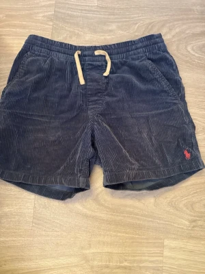 Blå manchester-shorts Polo Ralph Lauren - Kommer med lapp! Snygga blå manchester-shorts från Polo Ralph Lauren med dragsko i midjan och två sidofickor. Klassisk röd logga broderad på benet. Sköna och stilrena shorts i mjuk manchester som passar perfekt till sommaren.