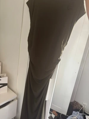Mörkgrön långklänning med drapering - Säljer en elegant mörkgrön långklänning med draperad effekt längs sidan. Klänningen har en enkel, ärmlös design och mjukt fallande tyg som ger en snygg siluett. Perfekt för dig som vill ha en stilren och trendig look.
