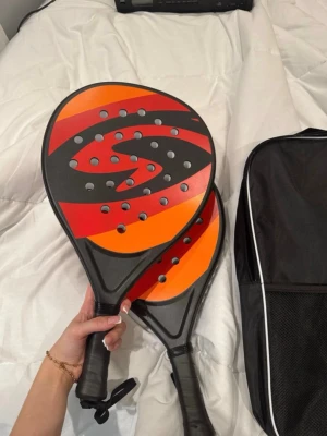  padelrack  - Säljer två padelracket i svart med röd och orange design, båda har hål i träffytan och svart grepp. Racketarna är märkta med 'Ultra Light' och 'Control'. En svart väska med vit kant och nätficka på sidan ingår, perfekt för att ta med till padelbanan. 