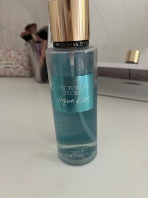 Victoria's Secret Aqua Kiss parfymmist - Fräsch parfymmist från Victoria's Secret i doften Aqua Kiss. Flaskan är cylinderformad med blått transparent innehåll och har en elegant guldfärgad kork. Rymmer 250 ml och ger en lätt, uppfriskande doft som känns modern och ungdomlig. Perfekt till sommaren. Den är använd men det är fortfarande ca3/4 kvar