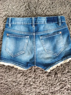 Blå jeansshorts med spetskant - Snygga blå jeansshorts med vit spetskant längst ner. Klassisk femficksmodell med knapp och dragkedja framtill. Shortsen har en tvättad look och är perfekta för varma dagar. Passar dig som gillar en söt men avslappnad stil. 