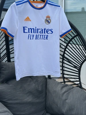 Real Madrid tröja 13-14y - Säljer en vit Real Madrid matchtröja från Adidas med blå och orange detaljer. Tröjan har klubbmärke och Adidas-logga på bröstet samt 'Emirates Fly Better' tryckt framtill. Tillverkad i lätt och ventilerande material, perfekt för fotboll eller träning.