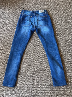 Blå slim fit jeans från Nudie - Snygga blå slim fit jeans med slitningar och tvättade detaljer på benen. Klassisk femficksmodell med normal midja och raka ben. Jeansen har kontrastsömmar och patch bak i midjan. Perfekta för en avslappnad och trendig look.