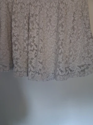 Vit kort kjol med spets och blommönster - Söt vit kort kjol med blommigt spetsmönster. Kjolen har en volangig form och är perfekt för dig som gillar romantisk och feminin stil. Materialet är tunt och luftigt, vilket gör den skön att bära under varmare dagar.