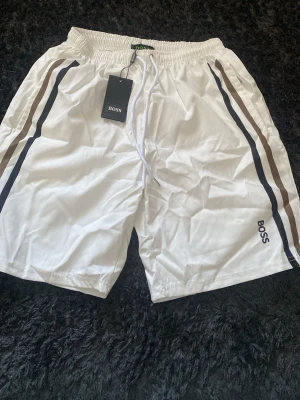 Vita shorts med ränder från BOSS - Snygga vita shorts från BOSS med svarta och bruna ränder längs sidorna. De har elastisk midja med snörning och logga på vänster ben. Perfekta för en avslappnad och sportig stil.