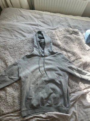 Grå basic hoodie med ficka - En enkel och stilren grå hoodie med huva och dragsnören. Hoodien har en stor magficka framtill och ribbade muddar vid ärmslut och nederkant. Perfekt för en avslappnad och chill look. Materialet känns mjukt och bekvämt, troligen bomullsblandning.