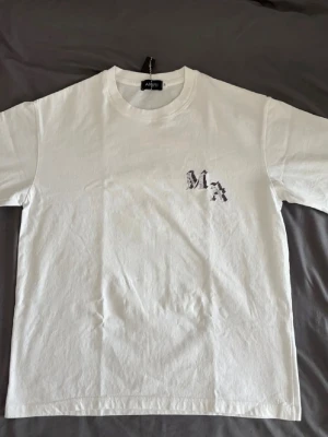 Vit t-shirt från Amiri  - Säljer en stilren vit t-shirt från Amiri i bomull med ett svart MA-tryck på bröstet. Klassisk passform och rund halsringning. Perfekt för dig som gillar minimalistisk design med en twist. 