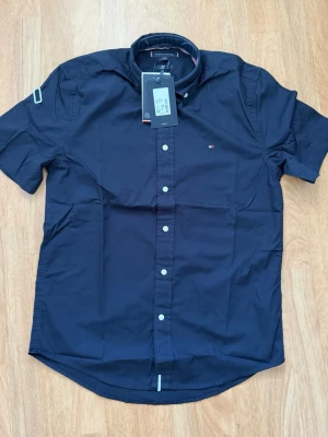 Mörkblå kortärmad skjorta Tommy Hilfiger - Snygg mörkblå kortärmad skjorta från Tommy Hilfiger med klassisk krage och vita knappar framtill. Regular fit-modell i bomull med liten logga på bröstet och diskreta detaljer vid kragen. Perfekt för en clean och stilren look.                                        Hade tänkt använda den själv men den blev tyvärr aldrig använd. Rensar garderoben och säljer därför vidare. Helt ny med alla lappar kvar.