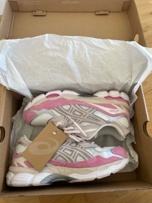 Asics Gel NYC - Snygga sneakers från Asics i vitt, rosa och beige. Skorna har mesh- och syntetpaneler för bra andningsförmåga och komfort. Klassisk Asics-logga på sidan och detaljer i rosa mockaimitation. Perfekta för en sportig och trendig look.