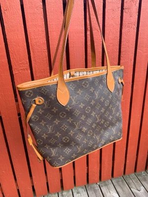 Monogram brun handväska med läderdetaljer - Snygg handväska i klassiskt brunt monogramtyg med ljusbruna läderhandtag och kanter. Väskan har öppen topp, långa axelband och justerbara sidoremmar. Perfekt för dig som gillar lyxig och tidlös design.