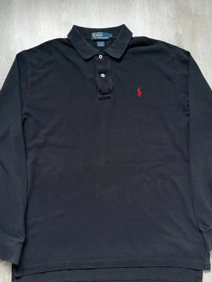Vintage Polo Ralph Lauren Långärmad Piké – Storlek L - Säljer en riktigt snygg och klassisk långärmad pikétröja från Polo by Ralph Lauren i storlek Large (L). Tröjan är i en stilren mörkblå/svart nyans tillverkad i kraftig 100% bomull pikékvalitet. Den har den ikoniskt broderade röda hästloggan på bröstet, knäppning i halsen och en helt slät baksida. Tröjan har en klassisk vintage-etikett och är i fint begagnat skick. Pris kan diskuteras. 