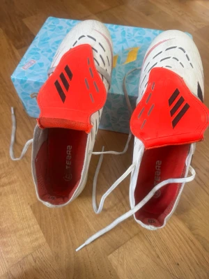 Vita och röda fotbollsskor - Säljer ett par vita fotbollsskor med röda detaljer och svarta ränder. Skorna har snörning och en modern design med ventilerande hål på ovandelen. Perfekta för dig som vill sticka ut på planen och få bra grepp. Skorna har en låg profil och är tillverkade i syntetmaterial.