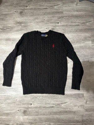 Svart kabelstickad tröja Polo Ralph Lauren - Kabelstickad svart tröja från Polo Ralph Lauren med rund halsringning och den klassiska röda logotypen broderad på bröstet. Långärmad modell i mjukt material som passar perfekt till höst och vinter, även till kalla sommarkvällar.Enkel och stilren design som funkar till det mesta. Säljer tyvärr detta klädesplagg då jag växt ur den.