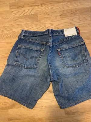 Blå denimshorts med fem fickor - Klassiska Levis 501 blå jeansshorts i denim med fem fickor och bälteshällor. Shortsens passform är normal och de har en knappgylf framtill. Perfekta för sommaren och ger en avslappnad look.