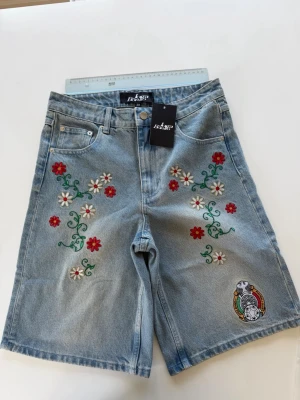 Joga Bonito Italia Jorts Light Wash S 28 Nya - Nya och helt oanvända Joga Bonito (Joga Bono) Italy Embroidered Jorts i ljusblått jeanstyg. Köpta för $80 men säljer pga fel storlek.  Storlek Small som motsvarar 28" i midjan, 78 cm runt om. Ben innersöm 28 cm, ben bredd 30 cm längst ned.   Detaljer: - Färg: Light wash / Ljusblå denim - 100% bomull, 14oz kraftig denim - Blommig brodyr längs benen + reworked Italy crest vid fållen - Skick: Nya med lapp, aldrig använda, aldrig tvättade