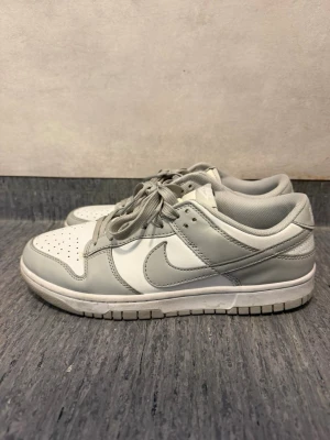 Nike vita och gråa sneakers - Snygga sneakers från Nike i vitt och ljusgrått. Skorna har klassisk låg siluett, perforerad tåbox och ikoniska swoosh-loggan på sidan. Tillverkade i läder med matchande grå skosnören och vit sula. Perfekta för en clean och stilren look.