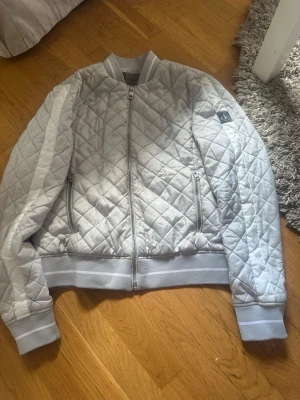 Ljusgrå quiltad bomberjacka CK - Snygg ljusgrå quiltad bomberjacka från Calvin Klein med dragkedja framtill, ribbade muddar och två fickor med dragkedja. Jackan har en klassisk bombermodell och en liten CK-logga på ärmen. Jackan är knappt använd, kanske använts 2 gånger. Den är i storlek M. 