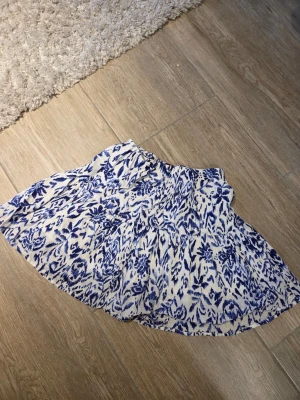 Vit och blå blommig kortkjol - Säljer en söt kortkjol med blått blommönster på vit botten. Kjolen har en luftig och flowy passform med volangdetaljer och resår i midjan. Perfekt för dig som gillar en avslappnad men trendig stil.