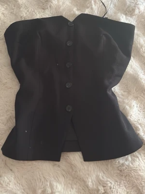 Svart korsettopp från BikBok - Snygg svart korsettopp från BikBok med knappar framtill. Toppen är axelbandslös och har en figurnära passform som framhäver midjan. Perfekt att styla med jeans eller kjol för en trendig look.