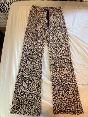 Leopardmönstrade pyjamasbyxor - Säljer ett par pyjamasbyxor med leopardmönster i beige och svart. Byxorna har raka ben och resår i midjan med svart dragsko för extra komfort. Perfekta för mysiga hemmakvällar.