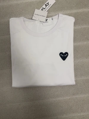 Vit t-shirt med svart hjärtdetalj - Säljer en vit t-shirt med rund hals och en svart hjärtformad patch med ögon på bröstet. T-shirten har en clean och stilren look, perfekt för dig som gillar minimalistisk design. Materialet ser ut att vara mjukt bomullstyg.
