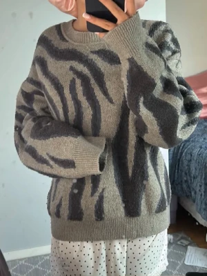  stickad tröja med zebramönster - Säljer en oversized stickad tröja med mörkgrått zebramönster. Tröjan har rund halsringning, långa ärmar och ribbade muddar vid ärmslut och nederkant. Perfekt för dig som gillar djurmönstrat och vill ha en mysig look. Lite nipprig vid ärmarna men inget som rikigt syns 