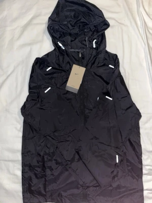 Svart Nike Millers med huva - Snygg svart vindjacka från Nike med reflekterande detaljer och huva. Jackan har dragkedja framtill och är tillverkad i lätt, prassligt material som passar perfekt för blåsiga dagar. Klassisk Nike-logga på bröstet och praktiska fickor.