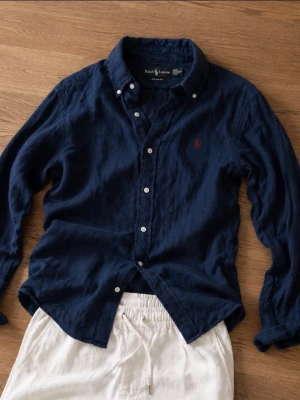 Mörkblå linneskjorta från Ralph Lauren - Säljer en mörkblå skjorta från Ralph Lauren i linne med klassisk krage och knappar framtill. Skjortan har lång ärm och den ikoniska röda logotypen broderad på bröstet. Perfekt för dig som gillar stilrena och tidlösa plagg.
