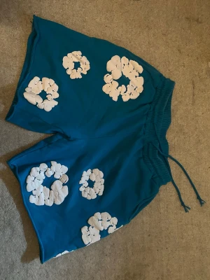 Blå shorts med vita blommor - Säljer ett par blå shorts med vita blommönster och snörning i midjan. Shortsen har en loose passform och är perfekta för sommaren. De har elastisk midja och är tillverkade i ett mjukt bomullstyg.