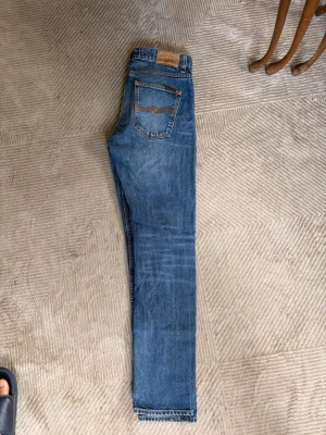 Nudie Lean Dean - Säljer ett par blå jeans med rak passform och fem fickor. Jeansen har en klassisk blå tvätt, orange sömmar och läderpatch bak i midjan. Perfekta för en avslappnad och stilren look. Materialet är denim i bomull.