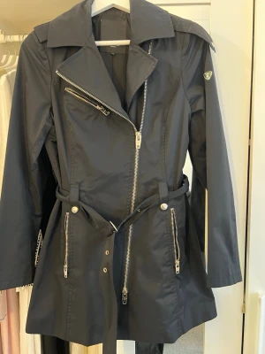 Svart trenchcoat med dragkedjor - Stilren svart trenchcoat med asymmetrisk dragkedja, bred krage och bälte i midjan. Jackan har flera silverfärgade dragkedjor och knappdetaljer vid fickorna samt på axlarna. Materialet ser ut att vara vattenavvisande och jackan har en figurnära passform.