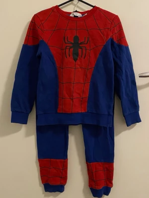 Spiderman pyjamas i rött och blått - Cool tvådelad Spiderman pyjamas med långärmad tröja och byxor. Tröjan är röd och blå med svart spindeltryck och nätmönster, medan byxorna är blå med röda detaljer och Spiderman-logga. Perfekt för Marvel-fans som vill sova i superhjältestil.