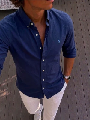 Marinblå skjorta från Ralph Lauren - Klassisk marinblå skjorta från Ralph Lauren med button-down krage och vita knappar. Skjortan har lång ärm och den ikoniska broderade loggan på bröstet. Tillverkad i bomull för en bekväm och stilren look.