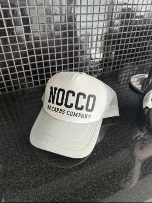 NOCCO keps - Snygg vit keps från NOCCO med svart text framtill. Kepsen har klassisk böjd skärm och meshmaterial baktill för extra ventilation. Justerbar storlek med plastspänne. Perfekt för en sportig och trendig look. Helt ny, aldrig använd. 