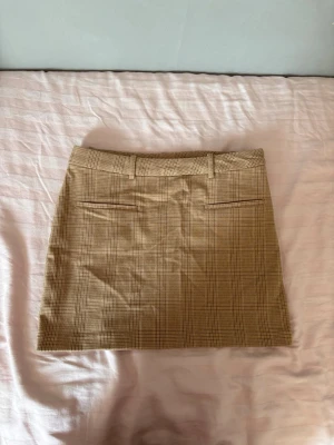 Beige rutig kortkjol med bälteshällor - Snygg beige kortkjol med rutigt mönster och bälteshällor i midjan. Kjolen har två dekorativa fickor bak och klassisk rak passform. Perfekt för en trendig och stilren look.