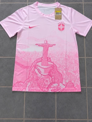 Rosa Brasil Nike t-shirt med tryck - Snygg rosa t-shirt från Nike med Brasilien CBF-logga och ikoniskt tryck av Kristusstatyn i Rio. T-shirten har rund hals, korta ärmar och är gjord i ett lätt, sportigt material som andas. Perfekt för dig som vill sticka ut med en unik och sportig look.