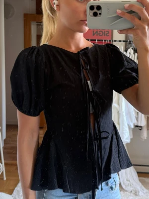 Svart blus med puffärm och knyt - Säljer en svart blus med puffärmar och knyt framtill. Blusen har en lätt utsvängd passform och är tillverkad i ett tunt, luftigt material. 