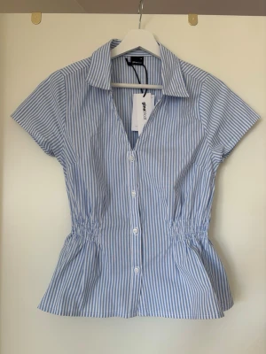 Blårandig kortärmad blus Gina Tricot - Snygg blårandig blus från Gina Tricot med korta ärmar och klassisk krage. Blusen har knäppning framtill och smock i midjan för en feminin siluett. Tillverkad av 50% ekologisk bomull. Perfekt till jeans eller kjol.