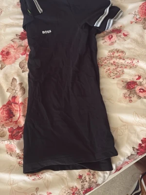 Svart pikétröja från BOSS - Säljer en svart pikétröja från BOSS med vit logga på bröstet. Tröjan har klassisk krage med knappar och vita detaljer på krage och ärmslut. Perfekt för en stilren och avslappnad look.