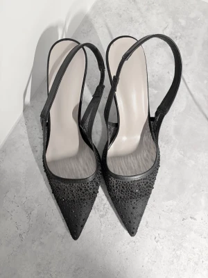 Svarta spetsiga pumps med strass - Säljer ett par svarta pumps med öppen häl och smal rem runt ankeln. Skorna har spetsig tå och är dekorerade med svarta strasstenar på ovandelen. Klacken är smal och elegant, och materialet ser ut att vara textil med glansig finish.