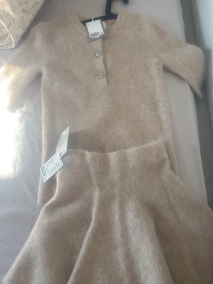 Beige stickat set med kjol och topp - Aldeig använd prislapp på Mysigt stickat set i beige med en topp som har tre glittriga knappar framtill och halvlånga ärmar. Kjolen är A-linjeformad och mjuk. Perfekt för dig som gillar en enkel men elegant look. Materialet känns fluffigt och bekvämt. Top storlek S kjol XS