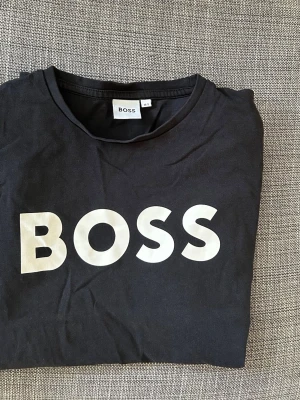 Svart BOSS t-shirt med stor logga. Strl 14 år - Snygg svart t-shirt från BOSS med stor vit logga framtill. Klassisk rund halsringning och korta ärmar. Perfekt för dig som gillar stilrena och trendiga plagg med tydlig branding. Storlek 14. S. 