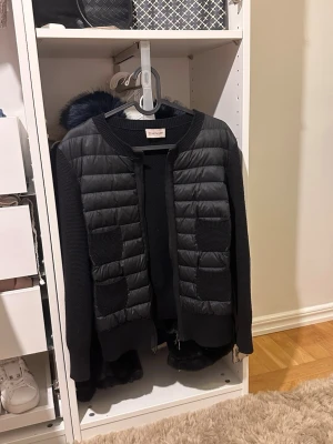 Svart cardiganjacka från Moncler - Snygg svart cardiganjacka från Moncler med quiltade partier framtill och stickade ärmar. Jackan har två framfickor, rund halsringning och dragkedja. Materialmix av vadderat tyg och stickat ger en modern look.