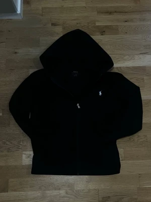 Svart hoodie med dragkedja - Svart hoodie med dragkedja och huva. Klassisk design med en liten vit logga på bröstet. Hoodien har långa ärmar och är perfekt för en avslappnad stil. Materialet ser ut att vara mjukt och bekvämt.