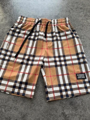 Rutig shorts med snörning - Säljer ett par rutiga shorts med klassiskt beige, svart, vit och röd färgkombination. Shortsen har snörning i midjan och en patch med texten 'BURBERRY LONDON ENGLAND' på benet. Perfekta för sommaren och har en avslappnad passform.
