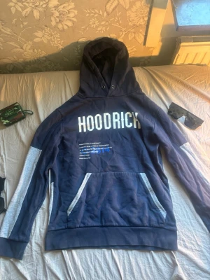 Marinblå HOODRICH hoodie med tryck - Snygg marinblå hoodie med stor vit HOODRICH-text på bröstet och inspirerande citat under. Grå detaljer längs ärmar och fickor. Klassisk känguruficka och huva med dragsko. Perfekt för en chill och street-inspirerad look.Kan gå ner något i pris vid en riktigt snabb och smidig affär! 