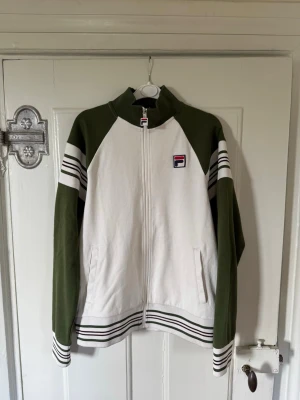 Fila fullzip  - Snygg ziptröja från Fila i vitt och olivgrönt med klassiska ränder på ärmar och nederkant. Tröjan har hög krage, dragkedja framtill och två fickor. Beställdes från 80scasualclassics och har bara använts ett fåtal gånger.