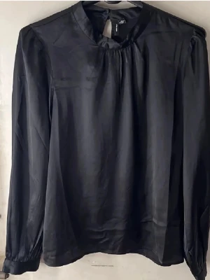 Svart satinblus med lång ärm - Stilren svart blus i glansigt satinmaterial med rundad halsringning och lätt puffade axlar. Blusen har långa ärmar med smala muddar och en knappdetalj i nacken. Perfekt för en trendig och elegant look.
