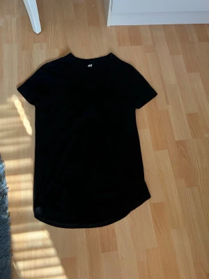 Svart lång t-shirt från H&M - En svart t-shirt från H&M i modellen Long Fit. T-shirten har rund halsringning och korta ärmar. Passformen är lite längre än vanligt vilket ger en avslappnad och modern look. Perfekt till jeans eller shorts.