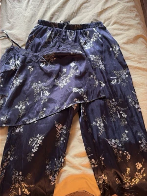 Mörkblå blommig satinpyjamas - Snygg tvådelad pyjamas i mörkblå satin med vita och ljusgröna blommor. Setet består av ett linne med spetsdetaljer och smala axelband samt ett par lösa byxor med resår i midjan. Perfekt för mysiga kvällar hemma. Skicka för mer info!💕